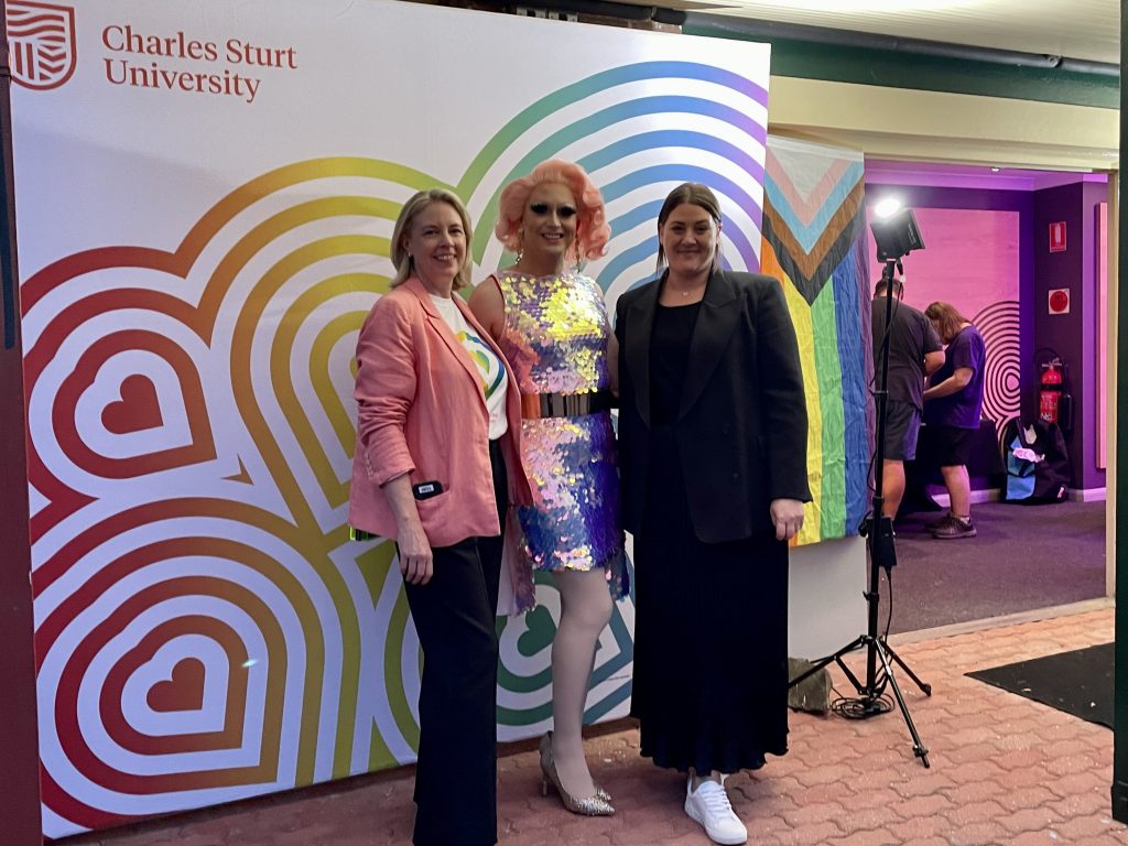 Drag queen Betty Confetti standing between Julia Andrews and Jac Underwood in front of a rainbow Charles Sturt pattern